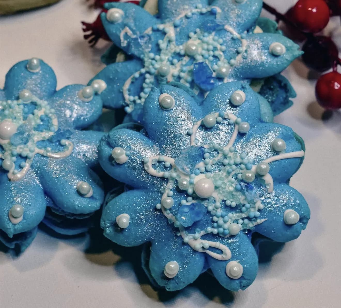 Sparkling blue snowflake-shaped macarons with white pearls and piped details alongside holly