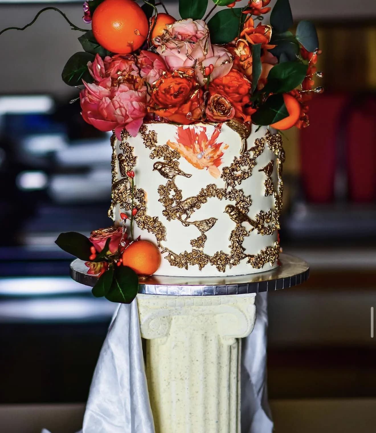 White cake with hand-painted gold filigree topped with oranges and coral roses on Greek column