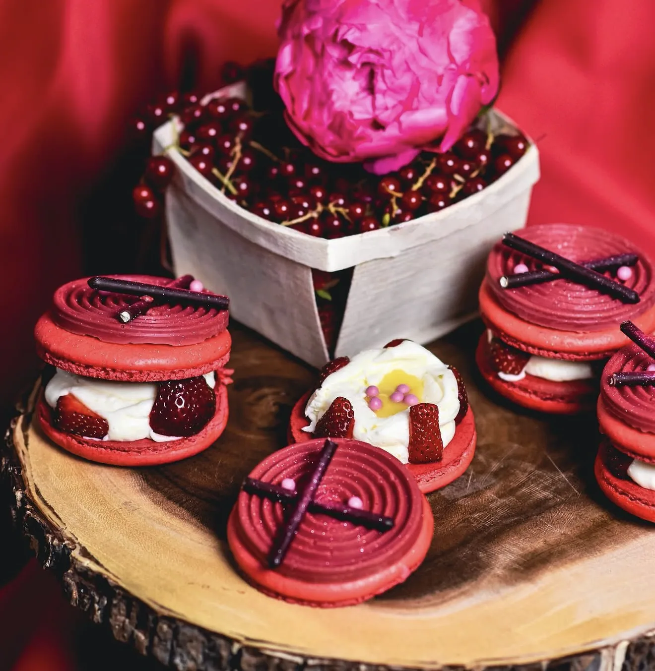 Spiral-piped red macarons with strawberries and pink peony atop rustic wood and red currants