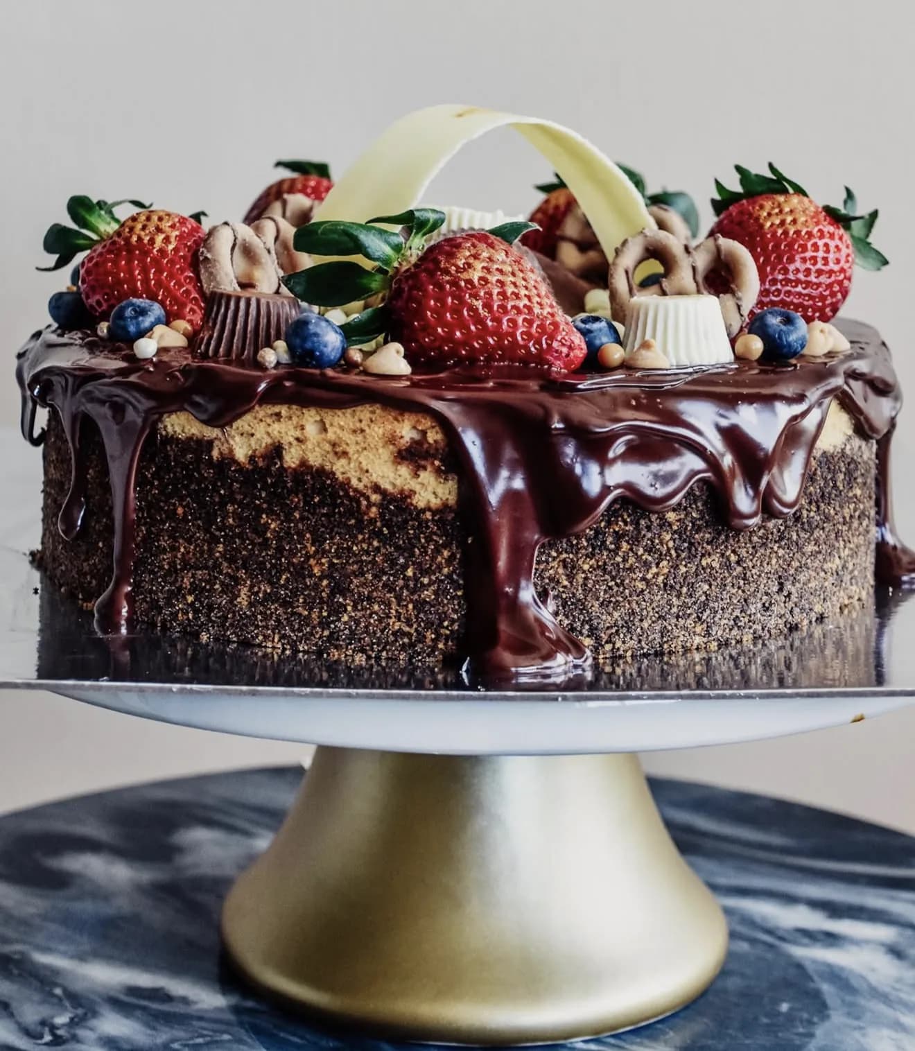 Side view chocolate cake with ganache drip and white chocolate arch atop fresh fruit on gold stand