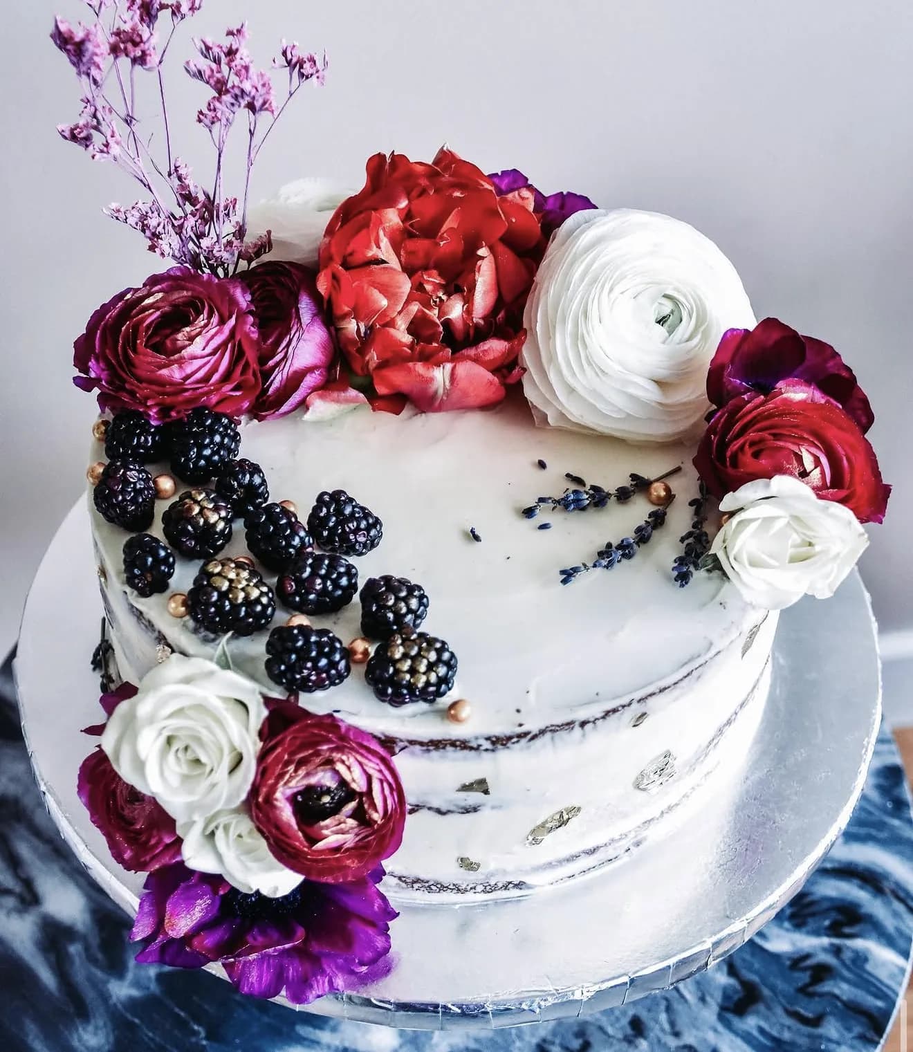 Semi-naked cake crowned with red peony blackberries lavender sprigs and burgundy ranunculus
