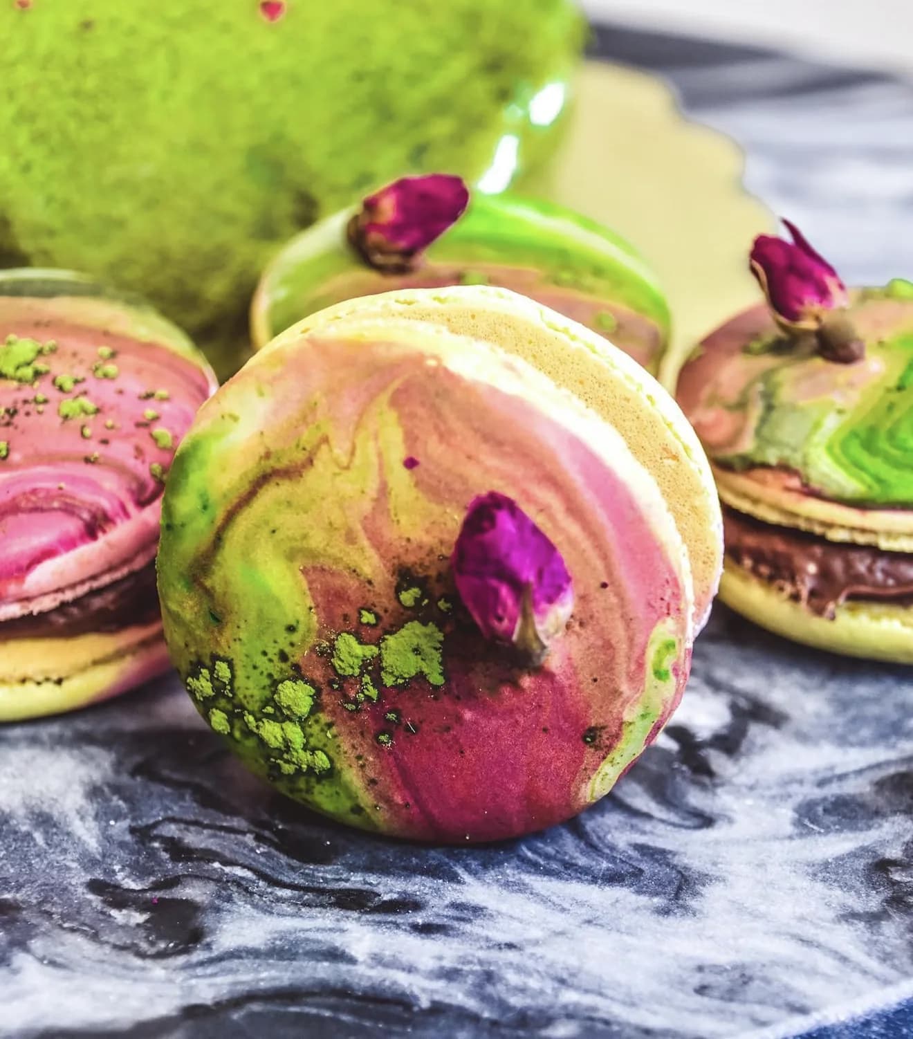Marbled pistachio and rose macarons with dried rosebud centers on blue marble surface
