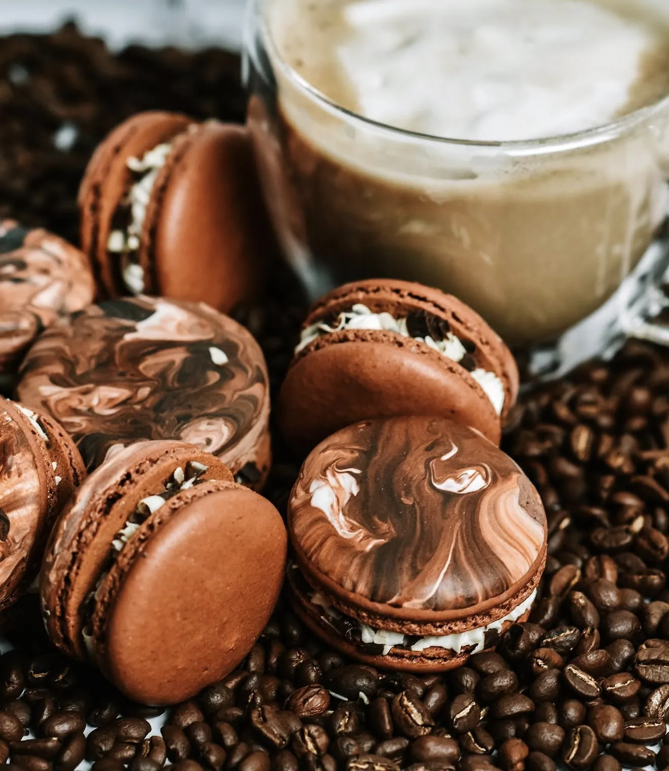 Side profile of mocha macarons stacked by frothy latte cup on coffee bean scattered surface