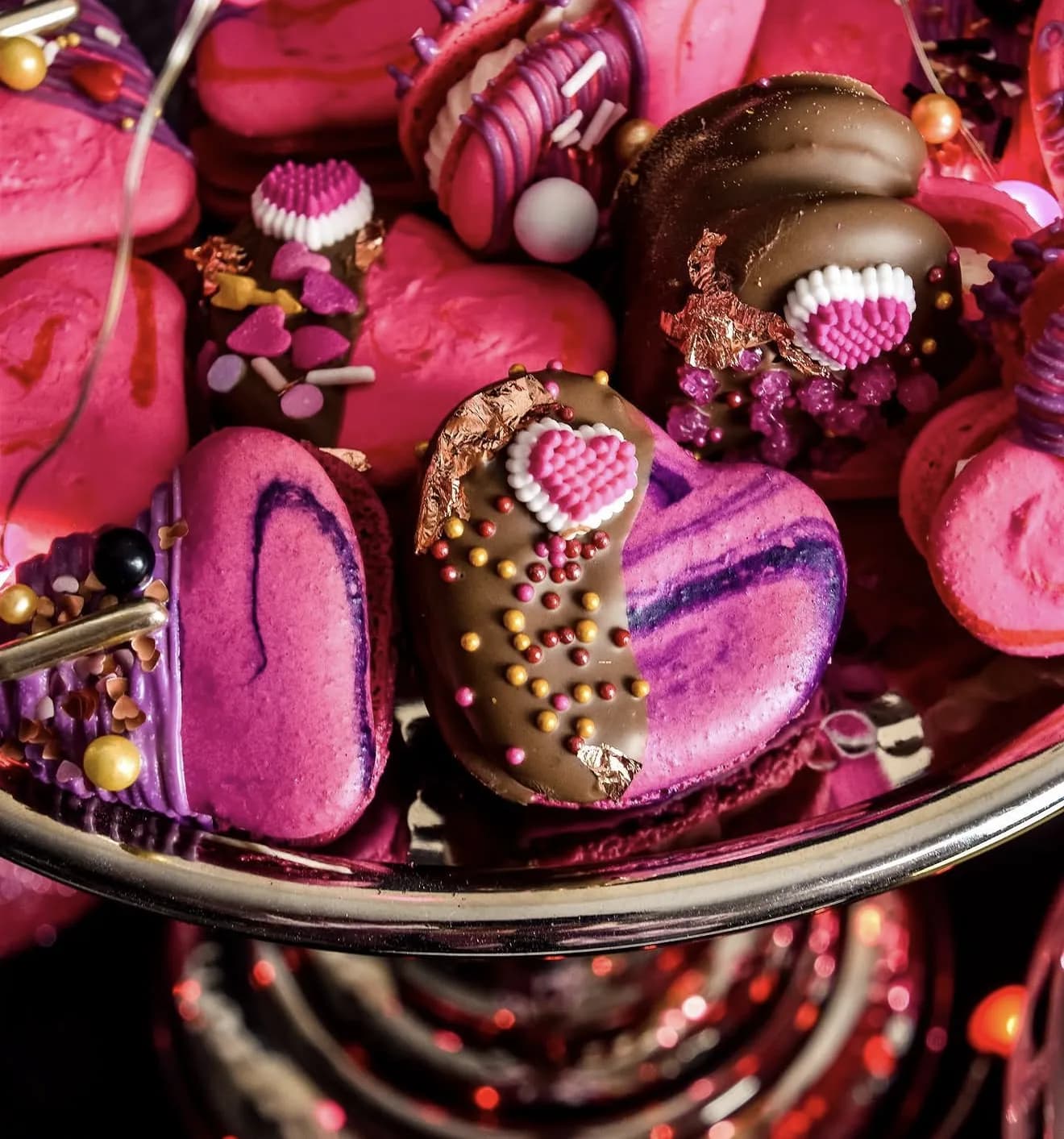 Bowl of Valentines pink and purple macarons with chocolate bonbons and sprinkles under fairy lights