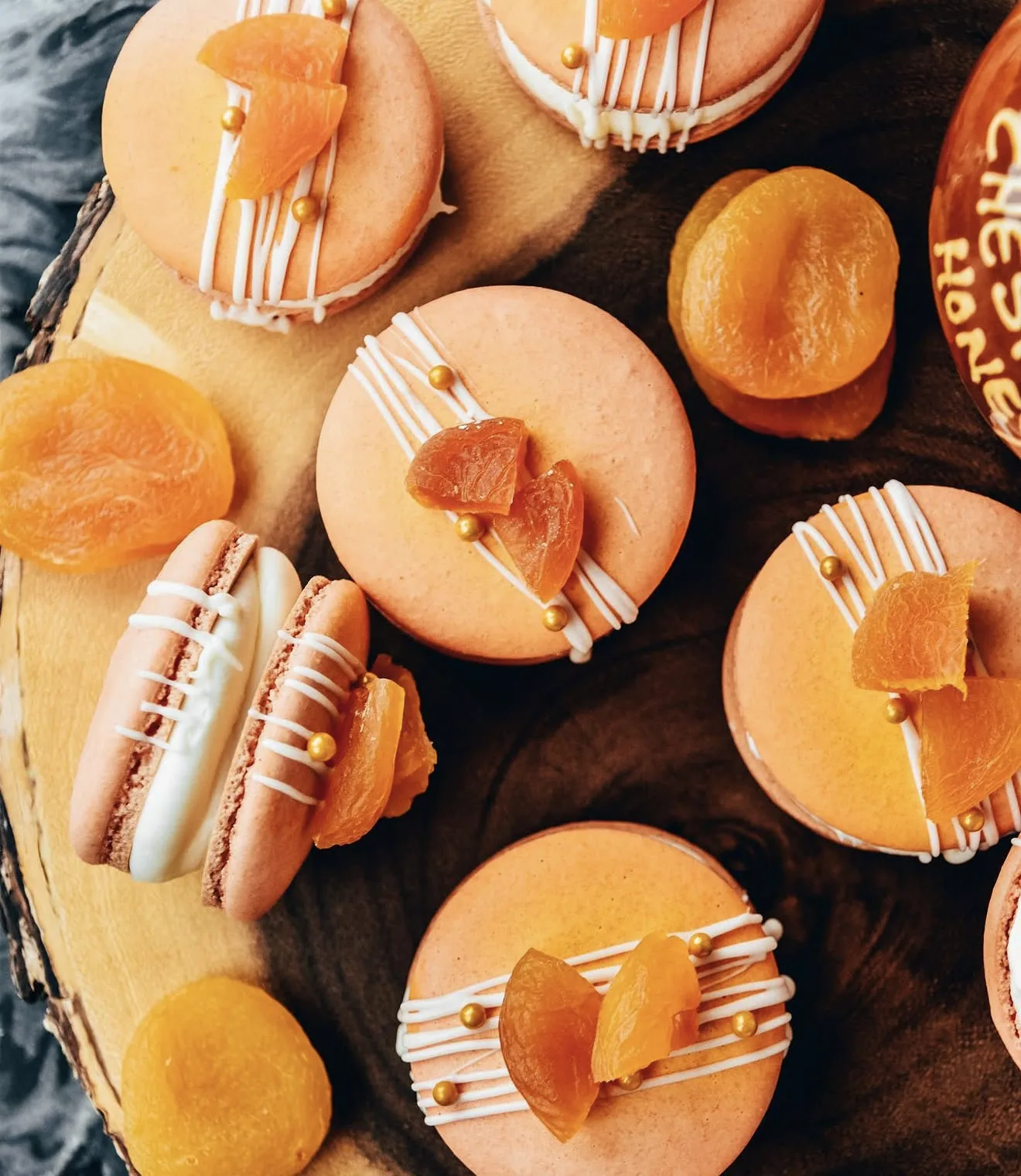 Apricot macarons around small chestnut honey pot with dried apricots on wood slab