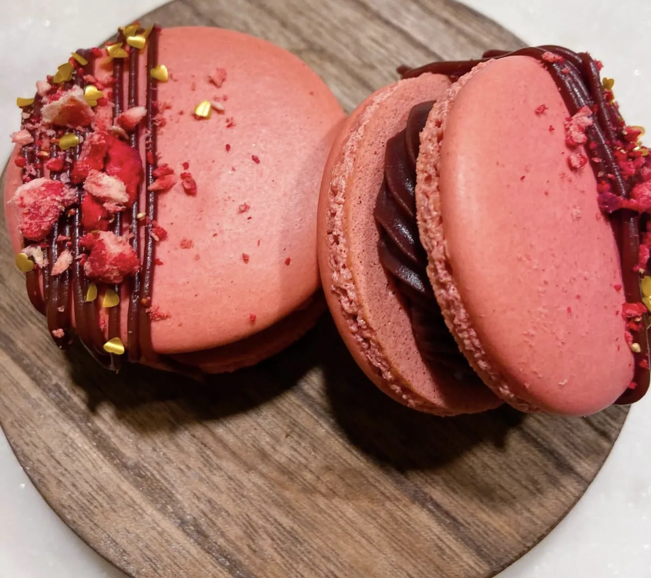 Side angle two chocolate macarons topped with twix bars and dripping caramel on wood stand