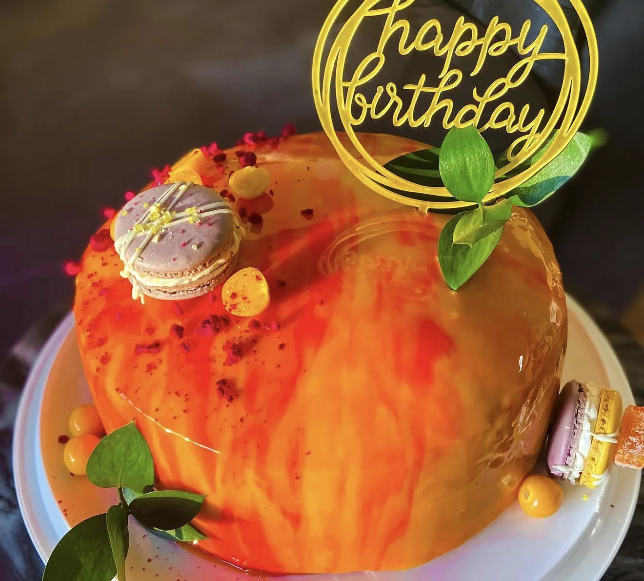 Wide angle orange mirror glaze cake with Happy Birthday hoop topper leaves and lilac macaron